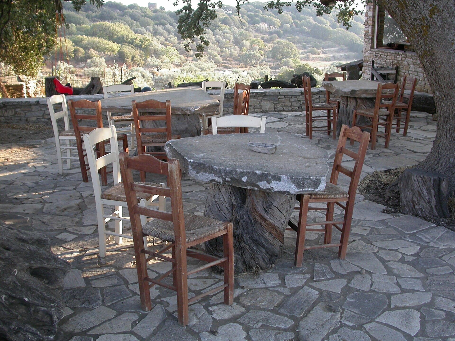 A stone terrace under a tree set with plain wooden chairs and round stone tables.